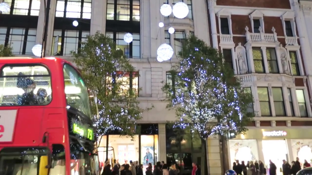 Red London bus passing beneath illuminated trees and Christmas globes on Oxford Street.