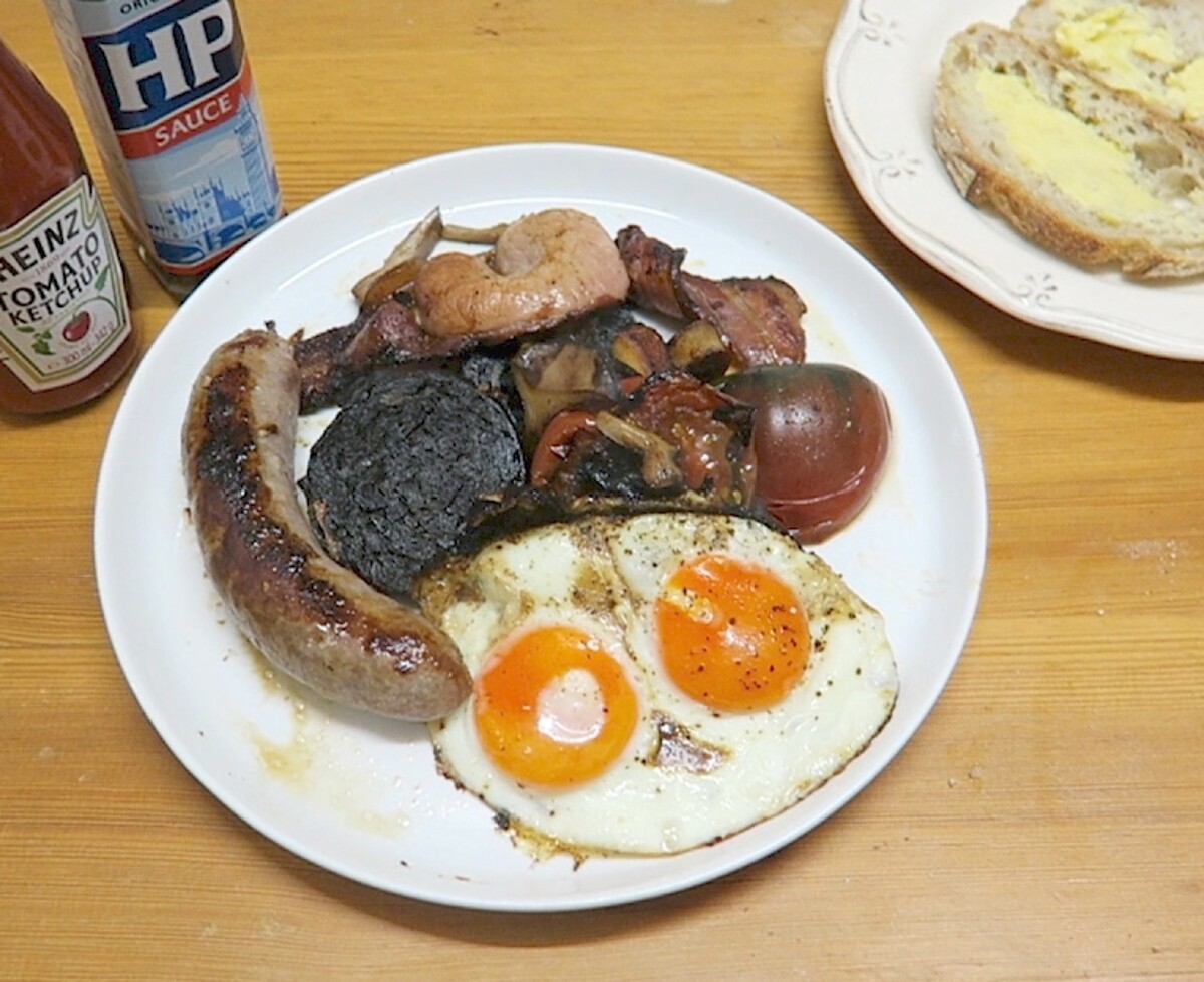 Close view of fried eggs, sausage, black pudding, bacon, mushrooms and tomato on the full English breakfast plate.
