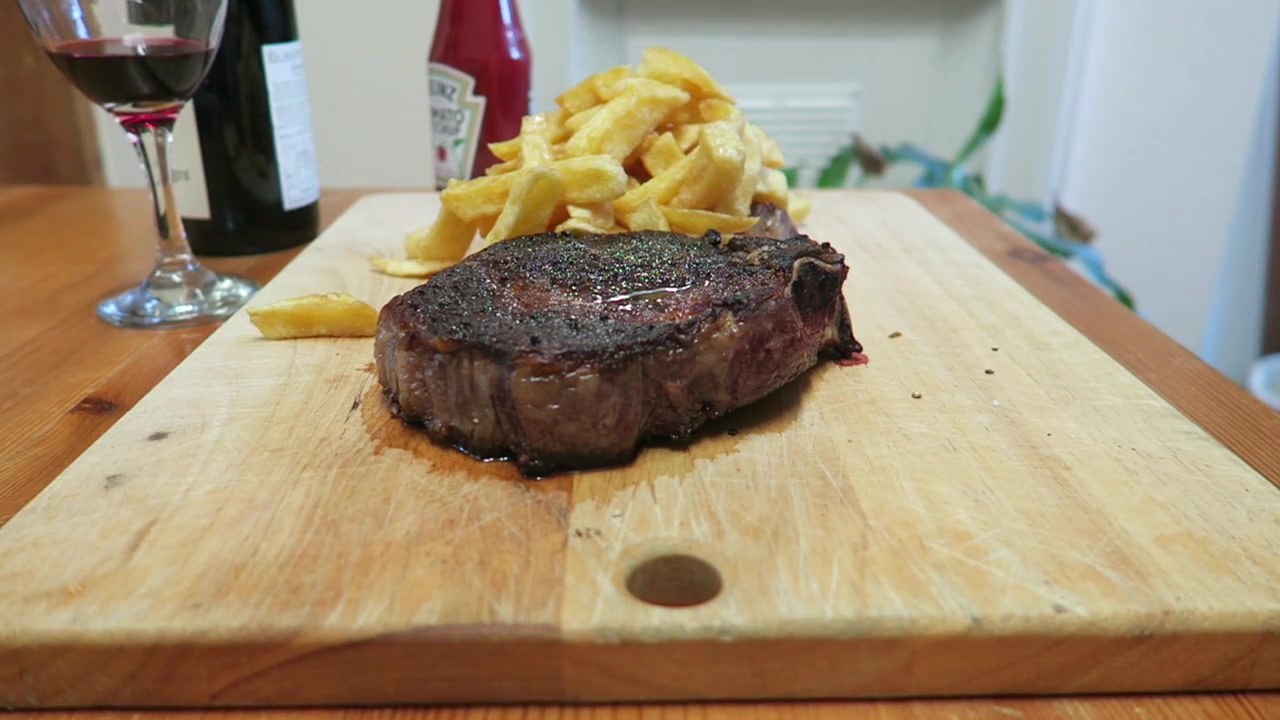 Cooked rib steak being sliced on a wooden board beside chips and red wine