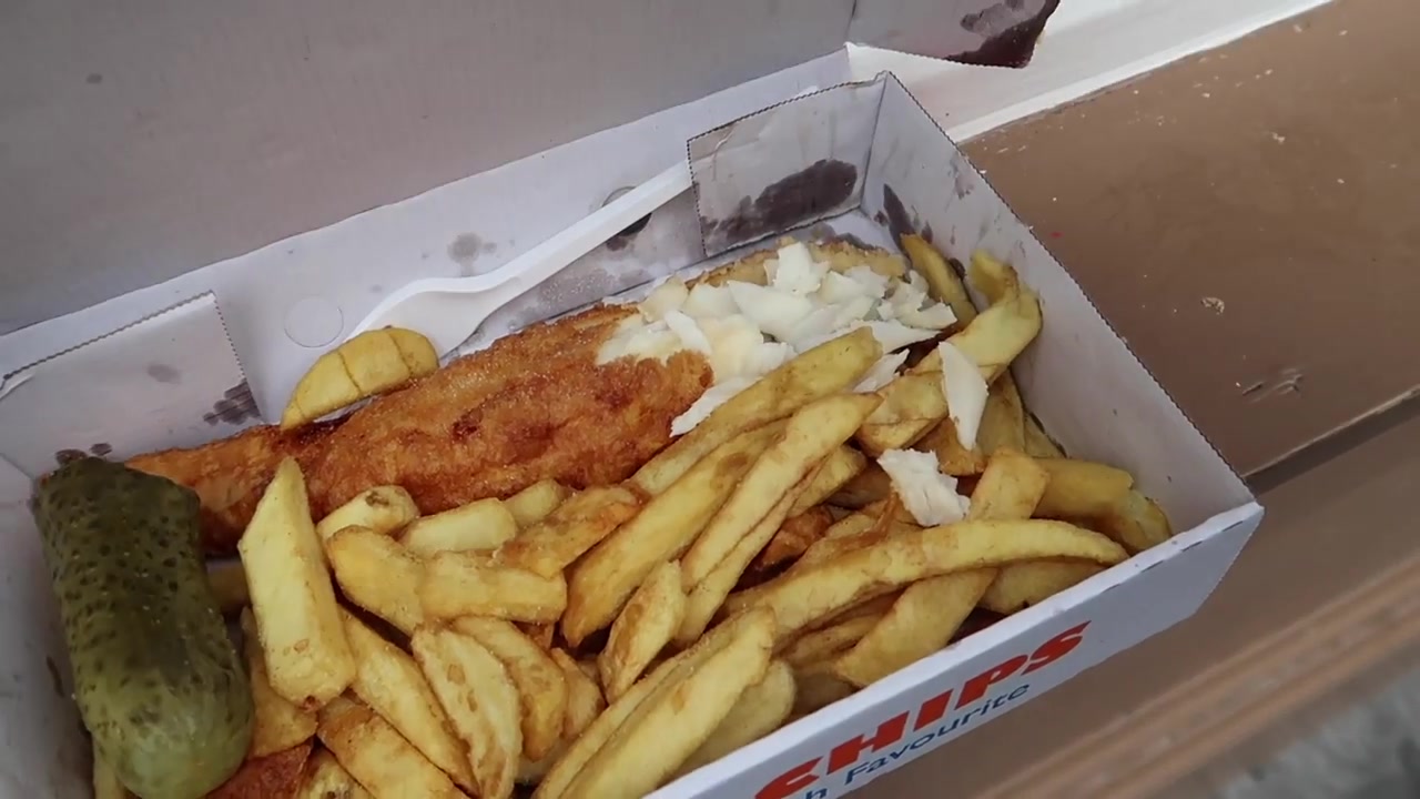 Close view of battered fish chips and pickled gherkin in a takeaway box