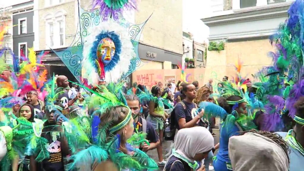 Masquerade performer in a vivid green and blue feathered costume at Notting Hill Carnival