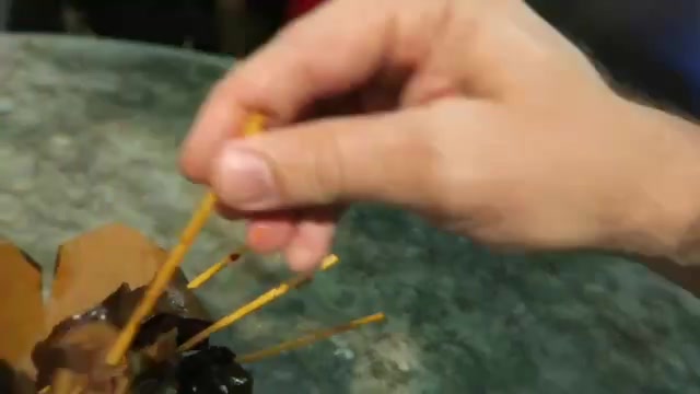A hand holding a skewer above a takeaway tub on a stone table