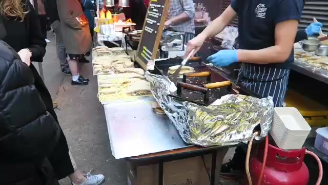 A trader cooking sandwiches on a griddle beside a market counter