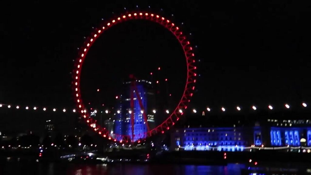 The London Eye glows red above the blue-lit riverside buildings.