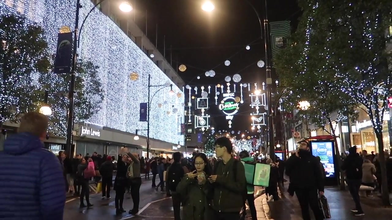 Shoppers beneath Christmas lights beside a brightly illuminated Oxford Street shopfront.