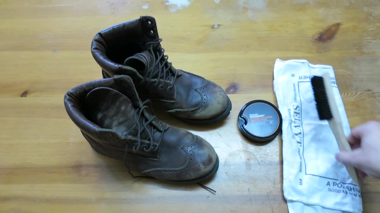 Worn leather boots with a Waximum tin, brush and cloth on a wooden table
