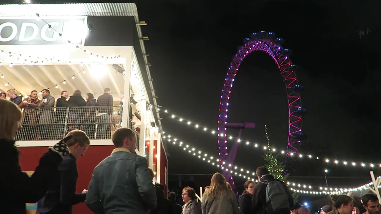 Christmas market visitors beneath festoon lights with the London Eye beyond.