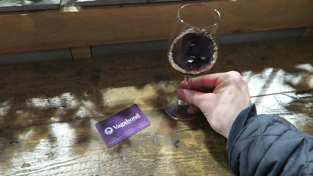A glass of red wine and a Vagabond card on a wooden table.