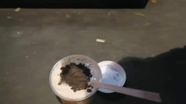 A takeaway cup of milk tea with a creamy topping and dark biscuit crumbs, filmed from above.